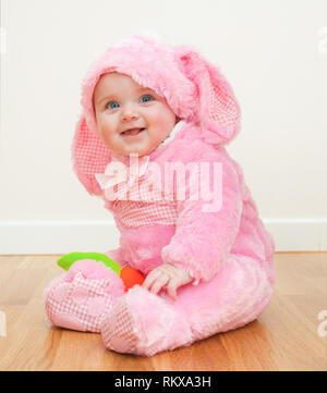 Little cute baby in a pink Bunny suit. The child sits on the wooden floor. Stock Photo