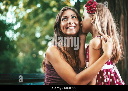 Beautiful mother and daughter are have fun at the park in spring day. Little girl is whispering something to her mother. Stock Photo
