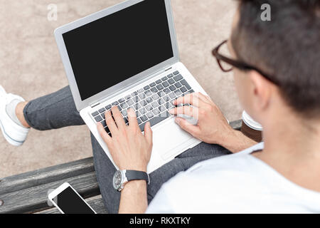 Young stylish man outdoors on the city street sitting on bench typing online message on laptop back view close-up Stock Photo