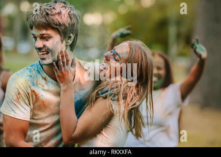 Smiling woman applying holi colour to the face of her boyfriend. Friends enjoying and having fun playing holi in a park. Stock Photo