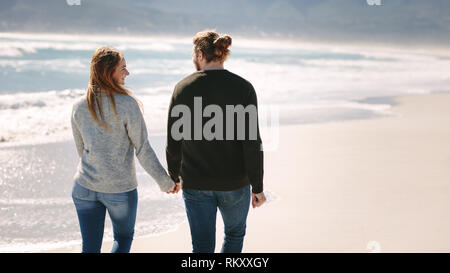 Rear view of romantic couple walking hand in hand on the beach. Man and woman taking a walk along the sea shore. Stock Photo