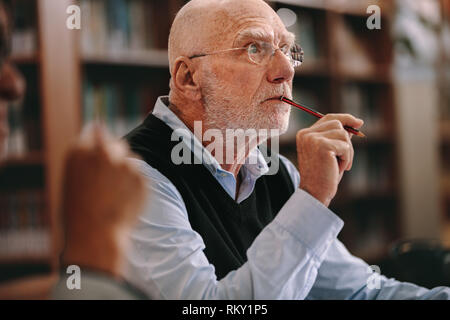 Elderly man sitting in classroom listening to a lecture with concentration. Senior man sitting in a library with pencil to his mouth and thinking. Stock Photo