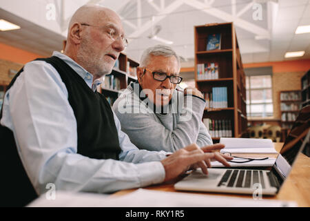 Side view of two elderly men working on laptop computer sitting in classroom. Senior men sitting in a library and learning their courses on a laptop. Stock Photo