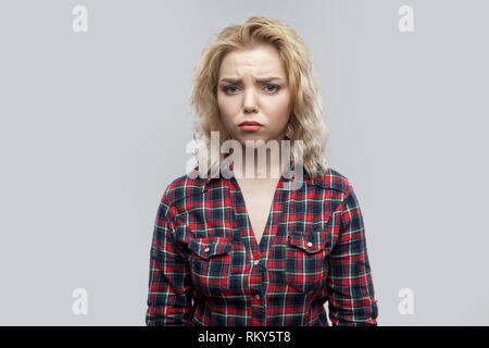 Portrait of unsatisfied beautiful blonde young woman in casual red checkered shirt standing and looking at camera with sad eyes. indoor studio shot, i Stock Photo