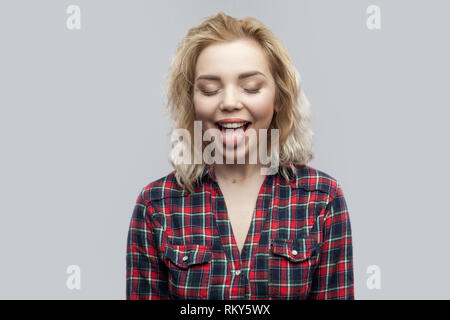 Portrait of funny beautiful blonde young woman in casual red checkered shirt standing with closed eyes and tongue out. indoor studio shot, isolated on Stock Photo