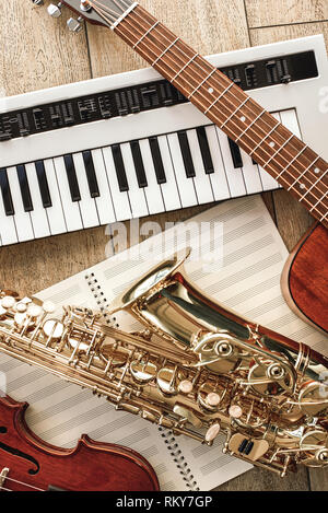 Vertical top view of different musical instruments: synthesizer, guitar, saxophone and violin lying on the sheets for music notes over wooden floor. Musical instruments. Music equipment Stock Photo