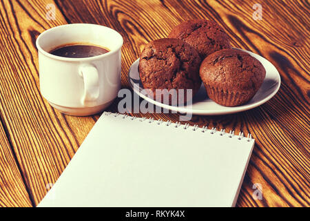 Several chocolate muffins with dark pastry with a cup of coffee and a notepad on a wooden table. Stock Photo
