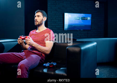 Man playing video games with gaming console and joy stick sitting on the couch in the dark room of the playing club Stock Photo