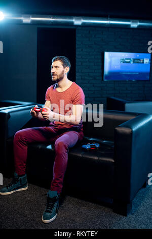 Man playing video games with gaming console and joy stick sitting on the couch in the dark room of the playing club Stock Photo