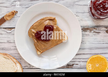 Top view of open face homemade peanut butter sandwich with dollop of heart shape strawberry jelly on oat bread, over a white rustic wooden table / bac Stock Photo