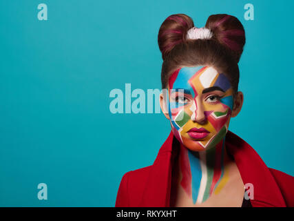 Portrait of funky girl with creative and colorful make up on face. Beautiful model wearing in red clothes, looking at camera, posing in studio. Woman has hairstyle bow, pink, plump lips. Stock Photo