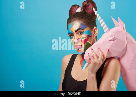Portrait of beautiful model posing, looking at camera. Charming woman holding hand on 3D paper unicorn head. Funky girl has pop art creative make up. Papercraft concept. Stock Photo