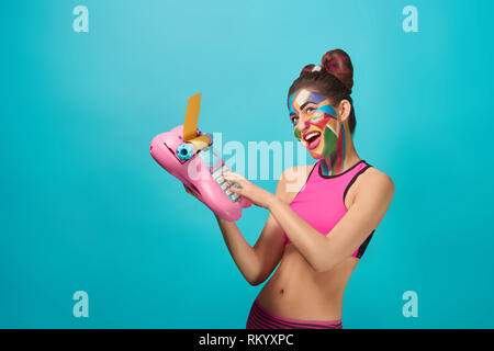 Happy and cheerful model looking up, smiling with opened mouth. Cool girl holding pink and blue toy typewriter. Funky, beautiful woman wearing pink top, has colorful pop art make up. Stock Photo