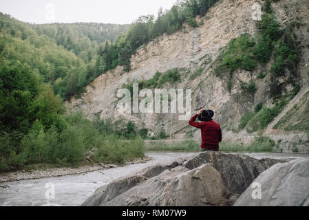 Young man in red jacket and black hat stands on a rock and enjoys mountain nature. Epic mountains on background. Concept outdoor nature lifestyle Stock Photo