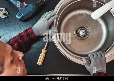 Inside view. Close-up of handsome plumber repairing sink in kitchen. Photo of metalic sink and handyman with screwdriver Stock Photo