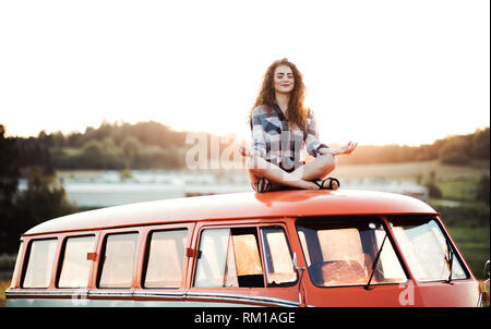 A young girl on a roadtrip through countryside, sitting on the roof of minivan doing yoga.