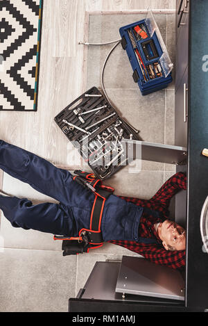 Tools case. Top view of seniour handyman repairing washbasin at the kitchen with professional equipment. He is lying on the floor Stock Photo