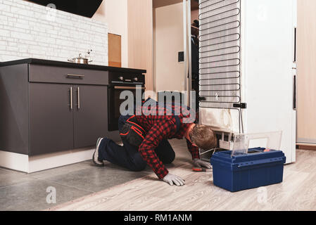 Ups, You have a trouble. Young Male Technician Checking Refrigerator With Screwdriver. He standing at knees Stock Photo
