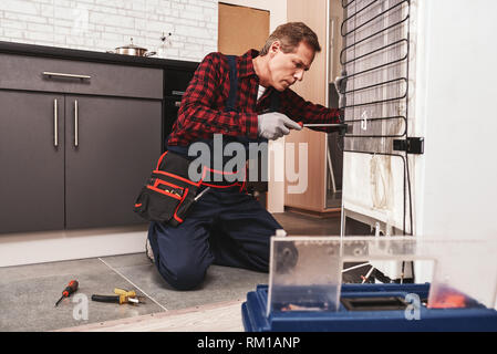 New refrigerator installation. Senior male technician checking refrigerator with screwdriver. He standing at knees near open toolbox. Side view Stock Photo