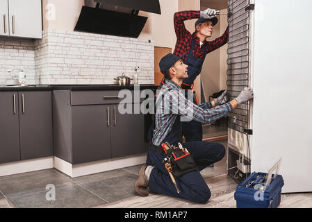 Two handymen with refrigerator. men mechanics checking refrigerator with screwdriver. They assist each other. Side view Stock Photo
