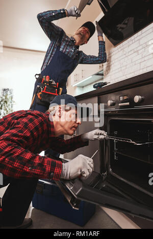 Productive together. Photo of two mechanics working at the kitchen. Senior specialist is repairing oven, young one is fixing exhaust hood Stock Photo