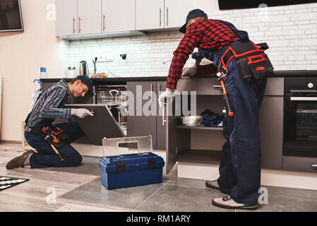 Learning from the best. Two men technician senior and young are sitting near dishwasher with screwdriver in kitchen with instruments Senior man teaches young one Stock Photo
