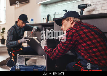 Quick and quality repairment. Two men technician senior and young are sitting near dishwasher with screwdriver in kitchen with instruments Senior man teaches young one Stock Photo
