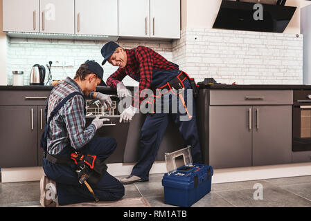 Take a pleasure from success. Two men technician senior and young are sitting near dishwasher with screwdriver in kitchen with instruments Senior man teaches young one Stock Photo
