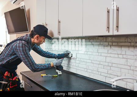 Electricity protection. Side view of young handyman setting new sockets with perforator at the kitchen Stock Photo