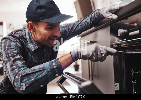 Don't delay with repair. Close-up of repairman examining oven with screwdriver in kitchen with tool case Stock Photo