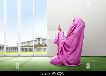 Young asian woman in pink veil sit in praying position on the carpet inside the room with glass door and mosque on the background Stock Photo