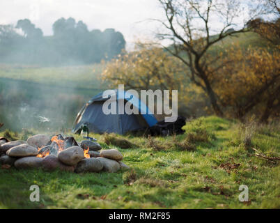 Smoking campfire in front of a dome tent pitched on the side of a riverbank. Stock Photo