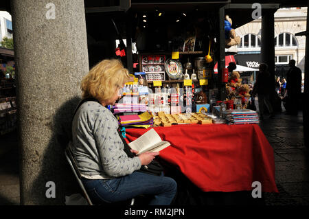 A senior woman spending a quiet moment reading a book at her market stall of tourist memorabilia, Covent Garden, London, England, UK Stock Photo