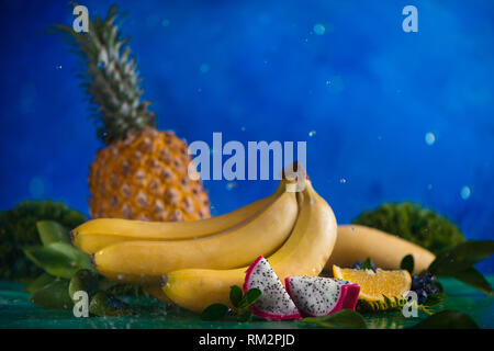 Pineapple, bananas, mango, dragon fruit with tropical leaves on a blue background with water drops and copy space Stock Photo