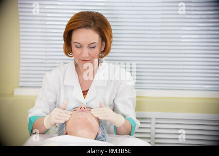 Doctor with red hair doing a facial massage to a young woman and talking to her during the procedure Stock Photo