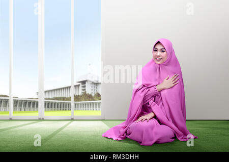 Smiling asian woman in pink veil sitting on carpet put her hand on chest and pray with all hearts inside the room with glass door and mosque on the ba Stock Photo