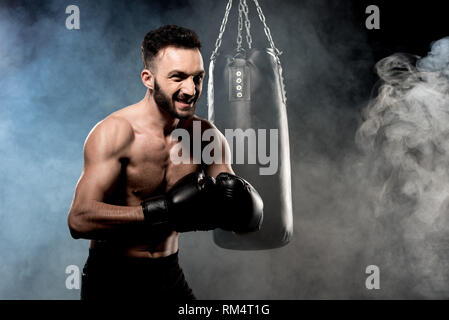 angry boxer standing in boxing pose near punching bag on black with smoke Stock Photo