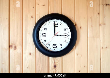 Big round wall clock with a black rim with arrows showing three o'clock hangs on brown wooden wall from vertical planks indoors on wooden background Stock Photo