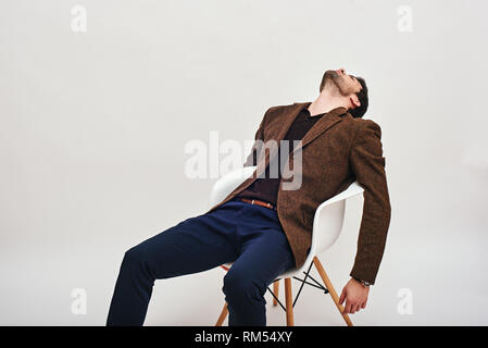 Portrait of stylish dark-haired man wearing blue trousers and brown jacket, sitting on a white chair, leaning back and looking at the ceiling isolated over white background Stock Photo