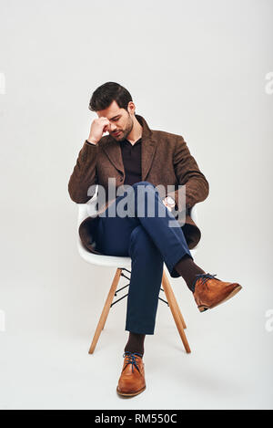 Full-length portrait of stylish dark-haired man wearing blue trousers and brown jacket, sitting on a white chair, exhausted after a hard day isolated over white background Stock Photo