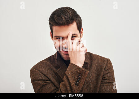 Portrait of stylish dark-haired man wearing brown jacket, touching his forehead, smiling and looking at camera isolated over white background Stock Photo