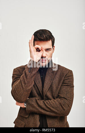 Portrait of a young handsome man wearing brown jacket, looking at camera and making OK sign, isolated over white background. Concept of success Stock Photo