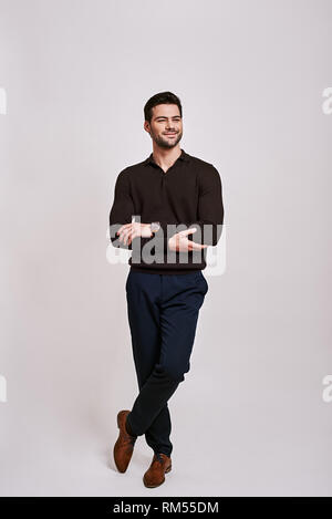 Full-length portrait of stylish dark-haired man wearing blue trousers and brown jumper, standing and looking away isolated over grey background Stock Photo