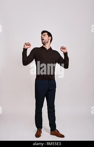 Full-length portrait of stylish dark-haired man wearing blue trousers and brown jumper, standing and yawning isolated over grey background Stock Photo