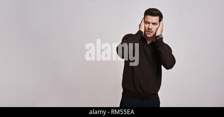 Portrait of surprised, shocked man with stubble and widened eyes and mouth in brown jumper touching his face with two hands isolated over grey background. Long shot Stock Photo
