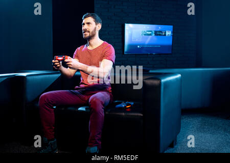 Man playing video games with gaming console and joy stick sitting on the couch in the dark room of the playing club Stock Photo