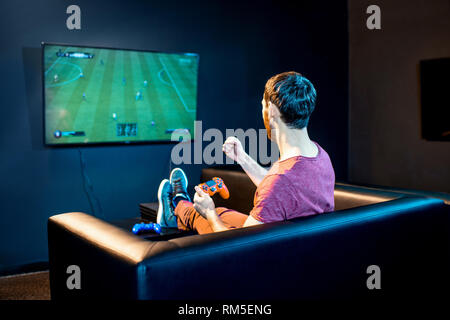 Man playing football game with gaming console sitting on the couch in front of the monitor at home or playing club Stock Photo