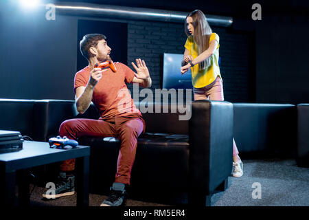 Young woman angry at her boyfriend playing video games with gaming console sitting on the couch at home or playing club Stock Photo