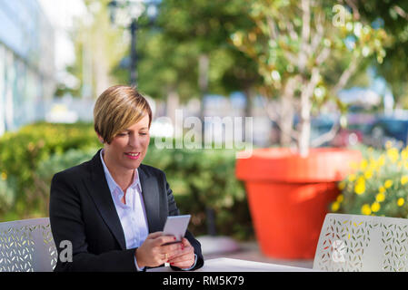 Front view of a elegant smiling business woman sitting on a chair in a coffee shop while using a mobile phone enjoying the conversation outdoors Stock Photo