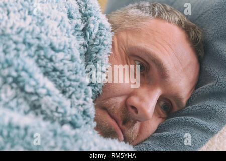 Mature man suffering from depression lies sadly covered with a blanket on his bed Stock Photo